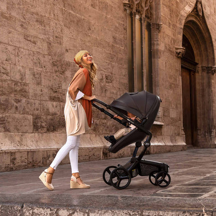 A parent walking the mima xari max Black stroller past a stone building with a child seated inside