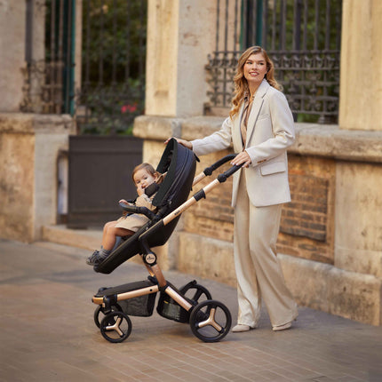 A toddler riding forward facing in the mima xari max on its Champagne chassis as a parent pushes along a sunlit street