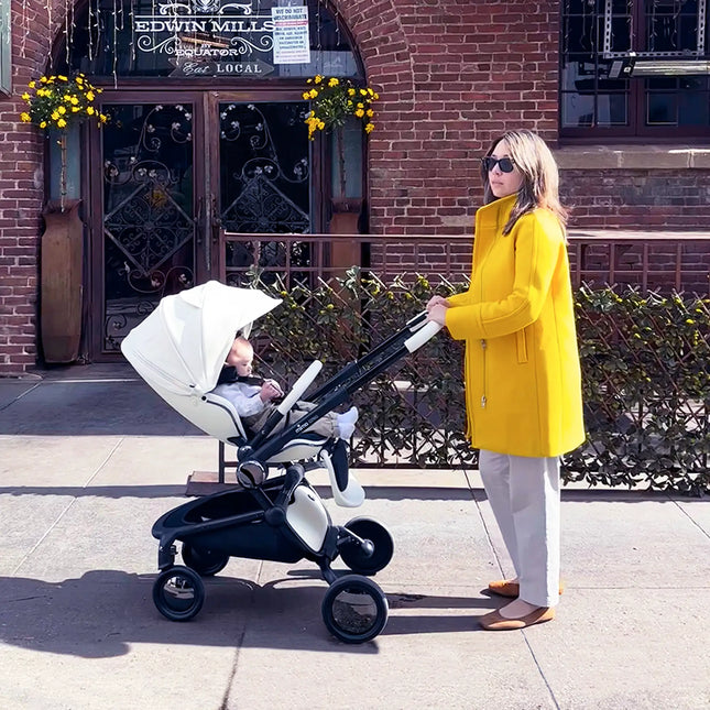 A parent in a yellow coat pushing the mima creo White stroller past a brick storefront