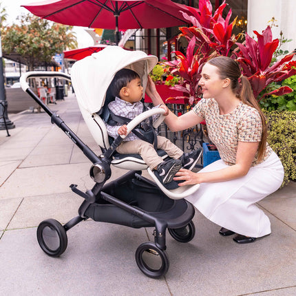 A parent playing with a toddler in the mima creo White city stroller under a red patio umbrella