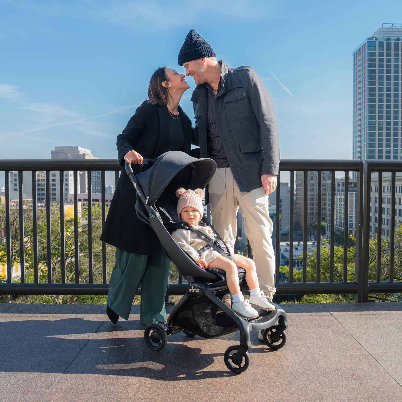 Parents on a rooftop city overlook with a toddler riding in the mima miro Black travel stroller