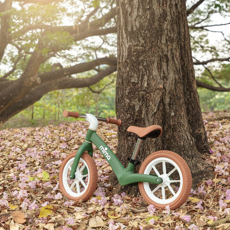 Green mima zoom lite balance bike leaning against a tree in fallen leaves