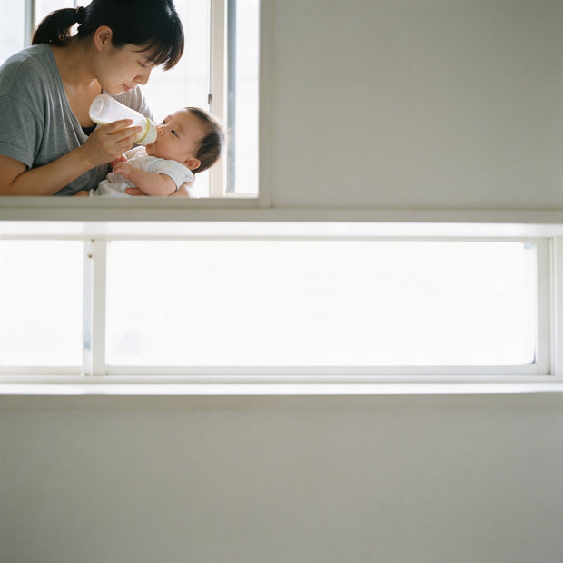 A mother bottle-feeds her baby by a window